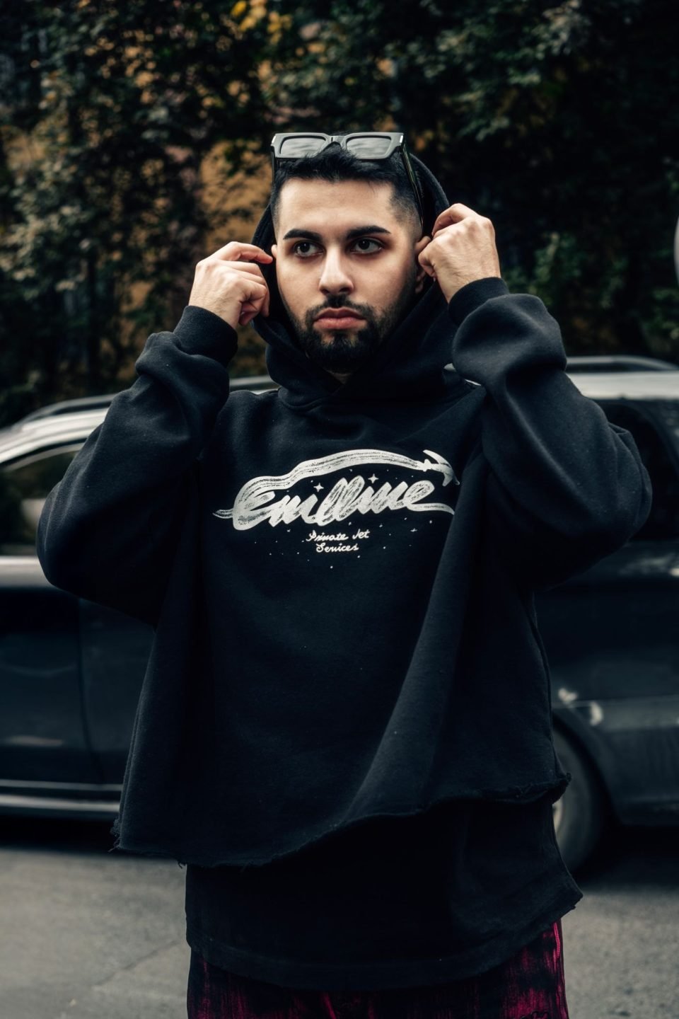 A young man in a black hoodie adjusting his glasses outdoors